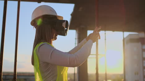 Woman-expert-engineer-Builder-in-VR-glasses-and-helmet-checks-the-progress-of-skyscraper-construction-moving-his-hands-at-sunset-visualizing-the-building-plan